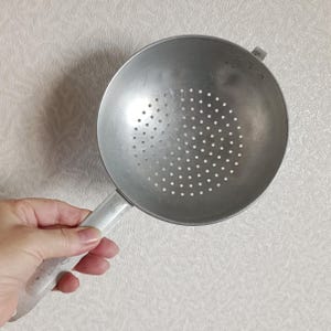 Puede incluir: Un colador de metal plateado con un mango y numerosos agujeros pequeños. El colador es redondo con un mango en un lado y un pequeño gancho en el otro. El mango es blanco.