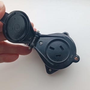 May include: Black electrical outlet with a hinged cover. The outlet has three slots and is designed to accept a three-prong plug. The cover is open, revealing the interior of the outlet. The outlet is mounted on a white surface.