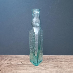 May include: A clear, light green glass bottle with a square base and a textured, pebbled surface. The bottle features a narrow, rounded neck that transitions into a wider, square-shaped body. The glass is translucent and reflects light.