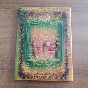 May include: A vintage journal with a detailed embossed cover. The cover features a cityscape design with spires and buildings, framed by ornate scrollwork. The color palette includes shades of green, brown, and gold, with the word "Vecriga" at the top.