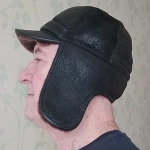 May include: A black leather aviator hat with ear flaps and a small brim. The hat has a textured surface and a soft, brown lining visible around the ear flaps. The hat is designed to provide warmth and protection from the elements.