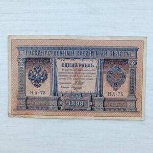 Op de afbeelding: Een rechthoekig Russisch bankbiljet van &eacute;&eacute;n roebel uit 1898 met ingewikkelde bruine en blauwe geometrische patronen. Het biljet toont de tekst "ГОСУДАРСТВЕННЫЙ КРЕДИТНЫЙ БИЛЕТЪ" en "ОДИНЪ РУБЛЬ" met een dubbelkoppige adelaar en een keizerlijk zegel.