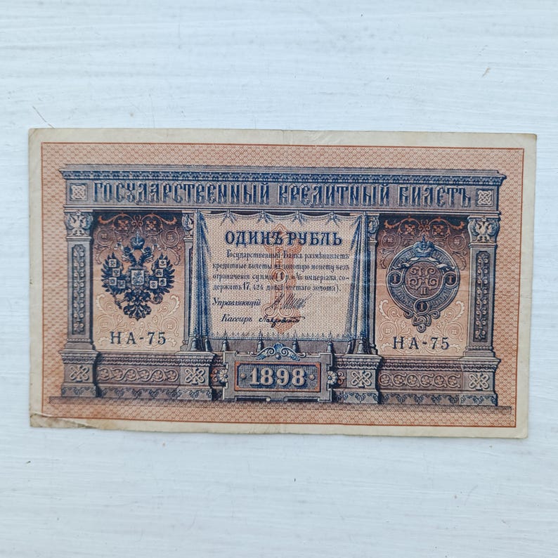 Op de afbeelding: Een rechthoekig Russisch bankbiljet van &eacute;&eacute;n roebel uit 1898 met ingewikkelde bruine en blauwe geometrische patronen. Het biljet toont de tekst "ГОСУДАРСТВЕННЫЙ КРЕДИТНЫЙ БИЛЕТЪ" en "ОДИНЪ РУБЛЬ" met een dubbelkoppige adelaar en een keizerlijk zegel.