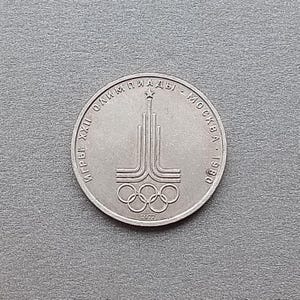 Op de afbeelding: Een zilveren munt met de Olympische ringen en een gestileerd torenontwerp. Er is Cyrillisch tekst zichtbaar, samen met het jaar 1980 en het nummer 1977. De munt ligt op een grijze ondergrond.