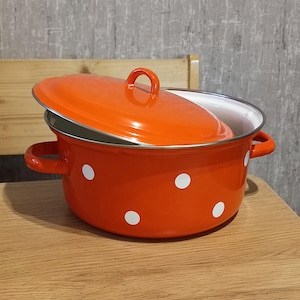 Op de afbeelding: Een oranje geëmailleerde pan met witte stippen en een bijpassend deksel. De pan heeft twee handvatten en staat op een houten oppervlak.