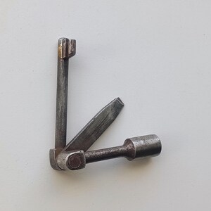 May include: A vintage, multi-tool wrench made of weathered metal. The tool features a socket, a slotted screwdriver, and a square-ended key. The metal has a worn, aged appearance, suggesting it is an antique or a well-used tool.