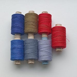 Op de afbeelding: Zes spoelen garen in verschillende kleuren, waaronder blauw, rood, grijs en bruin. De spoelen zijn gelabeld met de tekst "15K01", "15K01", "15K01", "15K01", "15K01" en "15K01".
