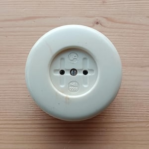 May include: Cream-colored electrical outlet with a circular design. The outlet features two holes and a central screw, with the text "~6A 220B" and a logo. The outlet is set against a light brown wooden surface.