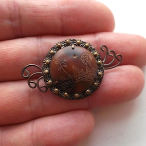 May include: A round, brown, carved, wooden brooch with a silver-toned metal frame and a floral design. The brooch has a delicate, wire-like design on the back.