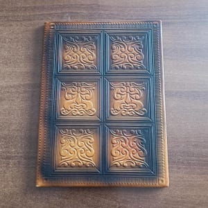May include: A rectangular, brown leather-bound journal with a decorative embossed design. The cover features a grid of six squares, each with an ornate, swirling pattern. The edges of the journal are also embossed with a repeating design.