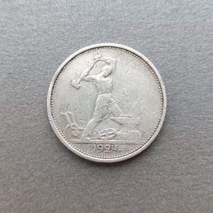 Op de afbeelding: Een zilverkleurige ronde munt met een reliëf van een figuur die een hamer op een aambeeld slaat. De figuur staat naast een ploegachtig werktuig. Het jaartal 1924 staat in reliëf onderaan de munt, die een geparelde rand heeft.