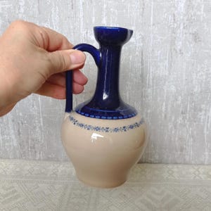May include: A ceramic pitcher featuring a glossy dark blue neck and handle, transitioning into a smooth beige body. A delicate blue floral pattern encircles the shoulder of the pitcher, separating the two colors with a decorative dotted border.