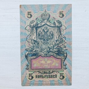 Op de afbeelding: Een vintage Russisch bankbiljet van vijf roebel met een centraal keizerlijk dubbelkoppig adelaarsschild, een kroon en sierlijke randen. Het papiergeld toont het cijfer 5 in de hoeken en cyrillische tekst in een centraal kader.