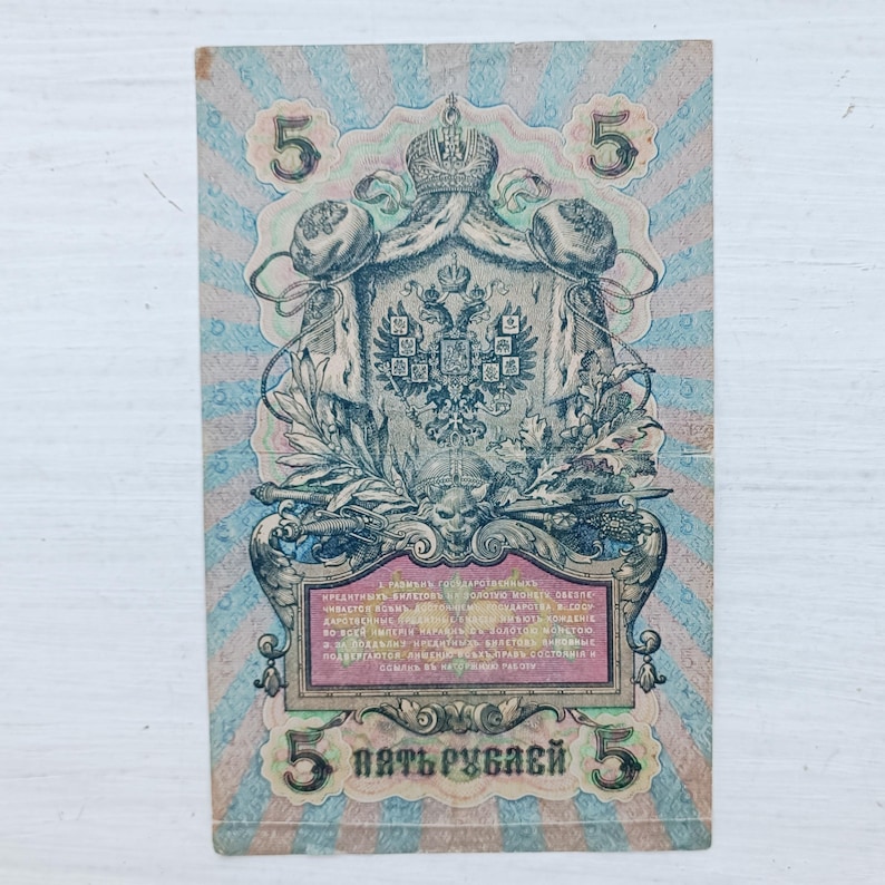 Op de afbeelding: Een vintage Russisch bankbiljet van vijf roebel met een centraal keizerlijk dubbelkoppig adelaarsschild, een kroon en sierlijke randen. Het papiergeld toont het cijfer 5 in de hoeken en cyrillische tekst in een centraal kader.