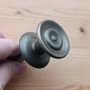 Puede incluir: Tirador de cajón de latón antiguo con un diseño circular escalonado. El herraje tiene un aspecto deslustrado y envejecido, lo que sugiere orígenes vintage. El tirador está montado sobre un fino vástago, adecuado para armarios o cajones.