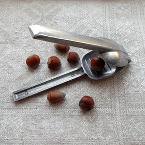 Puede incluir: Un cascanueces de plata con una manija de palanca y una base de metal. El cascanueces está rodeado de varias avellanas.