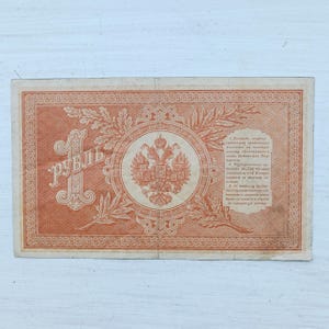 Op de afbeelding: Een rechthoekig Russisch bankbiljet van &eacute;&eacute;n roebel, gedrukt in oranje inkt. Het ontwerp bevat een groot cijfer &eacute;&eacute;n, het Cyrillische woord voor roebel, een centraal embleem met een dubbelkoppige adelaar, eiken- en laurierbladeren en tekst.
