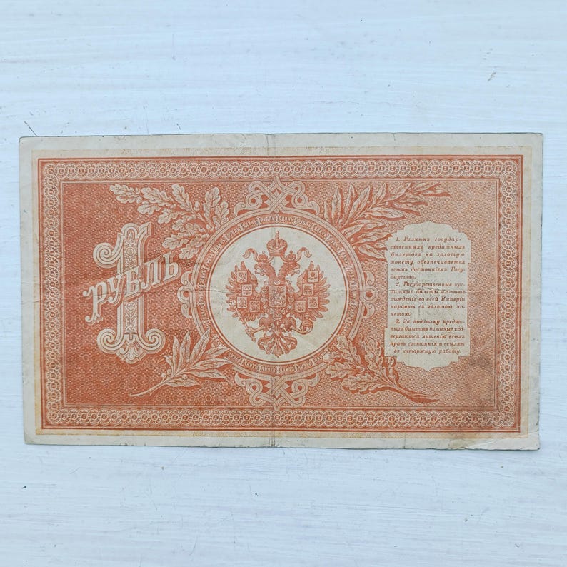 Op de afbeelding: Een rechthoekig Russisch bankbiljet van &eacute;&eacute;n roebel, gedrukt in oranje inkt. Het ontwerp bevat een groot cijfer &eacute;&eacute;n, het Cyrillische woord voor roebel, een centraal embleem met een dubbelkoppige adelaar, eiken- en laurierbladeren en tekst.