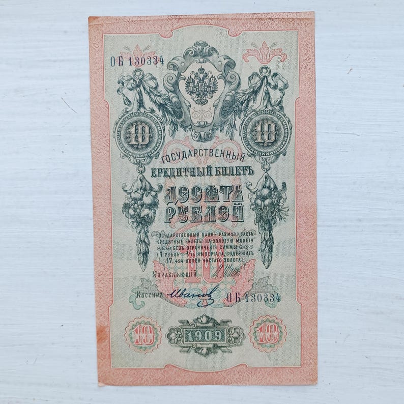 Op de afbeelding: Een vintage Russisch bankbiljet van tien roebel uit 1909 met ingewikkelde zwarte en groene krulmotieven, een centraal keizerlijk wapenschild en de tekst "ГОСУДАРСТВЕННЫЙ КРЕДИТНЫЙ БИЛЕТЪ ДЕСЯТЬ РУБЛЕЙ" op een beige achtergrond met een roze rand.