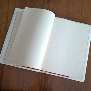 Puede incluir: Un cuaderno blanco con una encuadernación roja y páginas cuadriculadas. El cuaderno está abierto y acostado sobre una superficie de madera.
