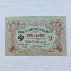 Op de afbeelding: Een Russisch bankbiljet van drie roebel uit 1905, voorzien van gedetailleerde groene, perzikkleurige en blauwe decoratieve randen. Het papiergeld toont het embleem van de tweekoppige adelaar, cyrillische tekst en serienummer BE 395964.