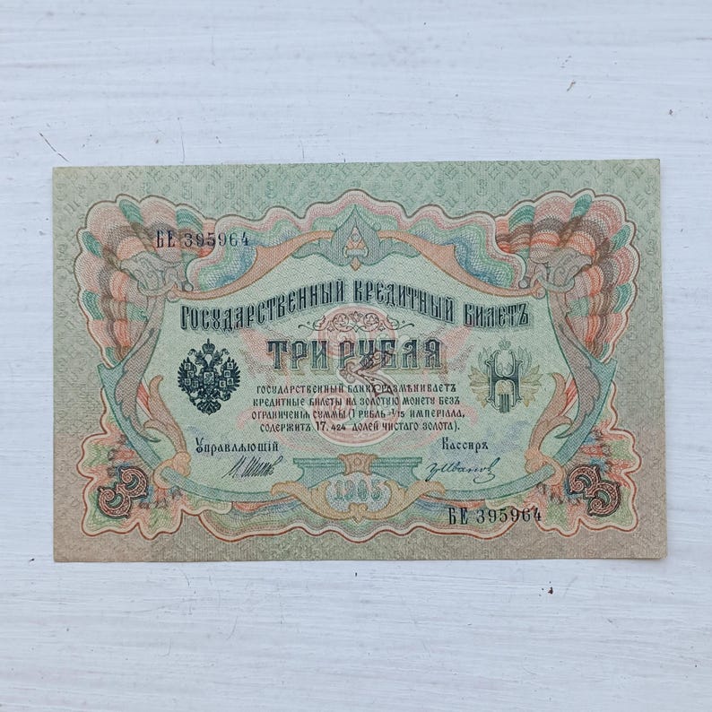 Op de afbeelding: Een Russisch bankbiljet van drie roebel uit 1905, voorzien van gedetailleerde groene, perzikkleurige en blauwe decoratieve randen. Het papiergeld toont het embleem van de tweekoppige adelaar, cyrillische tekst en serienummer BE 395964.