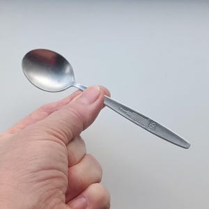 Peut inclure: Une cuillère en acier inoxydable avec un bol ovale et un manche décoratif. La poignée présente une série de lignes parallèles et un motif stylisé. La cuillère est tenue dans une main sur un fond uni.