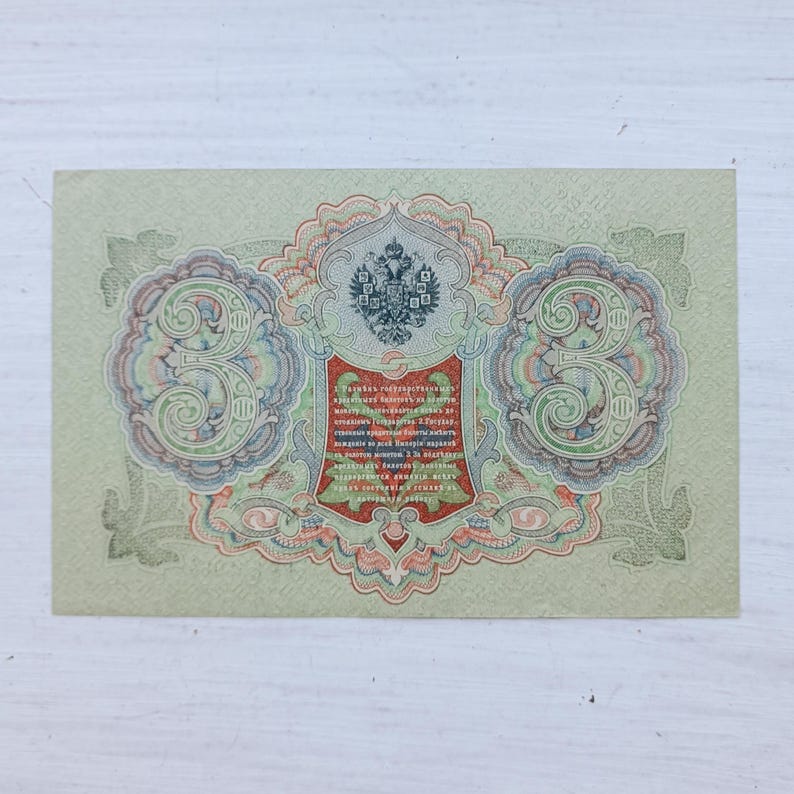 Op de afbeelding: Een vintage Russisch bankbiljet van drie roebel met een ingewikkeld guillochepatroon in groen, rood en blauw. Het ontwerp bevat een centraal embleem met een tweekoppige adelaar, grote cijfers drie aan beide kanten en cyrillische tekst in een rood schild.