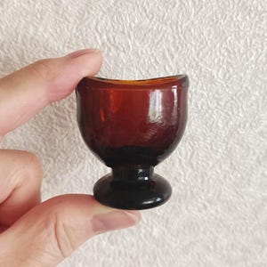 May include: A small, dark amber-colored glass egg cup featuring a rounded bowl shape, a short pedestal stem, and a circular base. The glass has a smooth, translucent finish with a curved rim designed to hold a single egg securely in place.