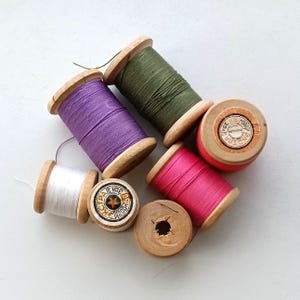May include: Six spools of thread in various colors, including white, purple, green, and pink. The spools are made of wood and have labels with text.