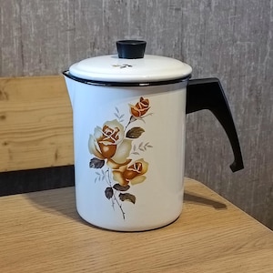 May include: White enamel pitcher with a black lid and handle. The pitcher features a floral design with yellow roses and green leaves.