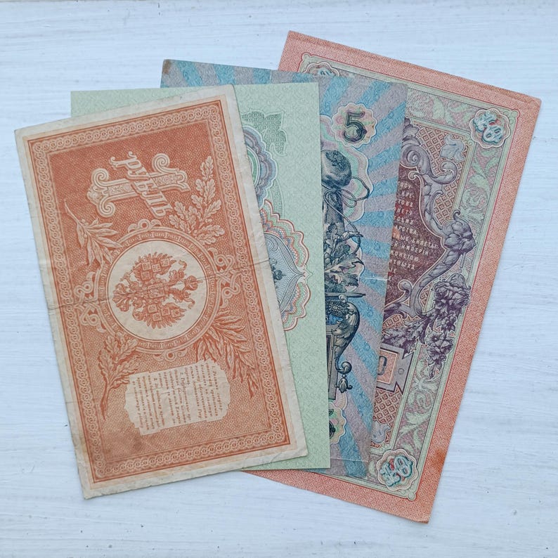 Op de afbeelding: Een verzameling van vier vintage papieren bankbiljetten die over elkaar heen liggen. De biljetten zijn voorzien van ingewikkelde decoratieve randen, cyrillische tekst, het cijfer 5 en een centraal dubbelkoppig adelaarsymbool in oranje, groen en blauw.