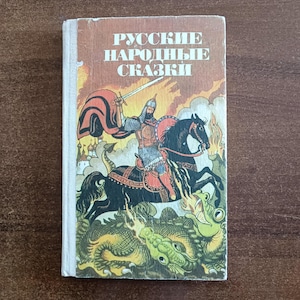 Pode incluir: Um livro vintage intitulado "Contos Populares Russos" com uma ilustração de capa de um cavaleiro a cavalo lutando contra um dragão. A capa apresenta um fundo castanho com texto em russo e uma ilustração colorida.