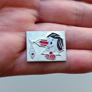 Op de afbeelding: Een zilveren metalen pin met een rood en zwart cartoonfiguur dat een vis vasthoudt. Het figuur draagt een rode hoed en een rood-wit gestreept shirt. De vis is rood en wit.