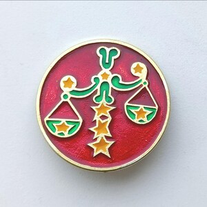 Könnte beinhalten: Eine kreisförmige Emaille-Anstecknadel mit leuchtend rotem Hintergrund und goldfarbenem Rand. Das Design zeigt eine zentrale vertikale Säule aus vier goldenen Sternen, flankiert von zwei grünen waagenartigen Strukturen mit je einem Stern.