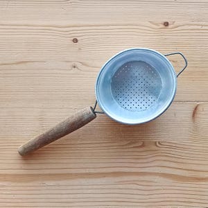 Könnte beinhalten: Ein silbernes Sieb mit einem Holzgriff. Das Sieb hat eine runde Schüssel mit einem perforierten Boden und einem Drahtbügelgriff.