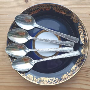 Könnte beinhalten: Vier Edelstahllöffel mit geprägten Blumenmustern auf den Griffen, angeordnet auf einer kreisförmigen, dunkelblauen Untertasse mit goldfarbenem Zierrand und einem weißen Mittelkreis, die eine reflektierende metallische Oberfläche aufweisen.