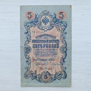 Op de afbeelding: Een vintage Russisch bankbiljet van vijf roebel uit 1909 met gedetailleerde blauwe en perzikkleurige decoratieve krullen, het keizerlijke dubbelkoppige adelaarsymbool en cyrillische tekst. Het biljet toont het serienummer TI 977293.