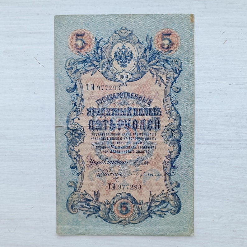 Op de afbeelding: Een vintage Russisch bankbiljet van vijf roebel uit 1909 met gedetailleerde blauwe en perzikkleurige decoratieve krullen, het keizerlijke dubbelkoppige adelaarsymbool en cyrillische tekst. Het biljet toont het serienummer TI 977293.