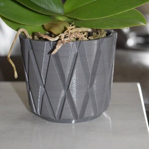 Op de afbeelding: Een grijze keramische pot met een ruitpatroon, met daarin een groene orchidee met bruine wortels.