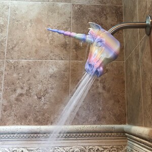 May include: A unicorn shower head with a rainbow design. The shower head is attached to a silver shower arm and is spraying water.