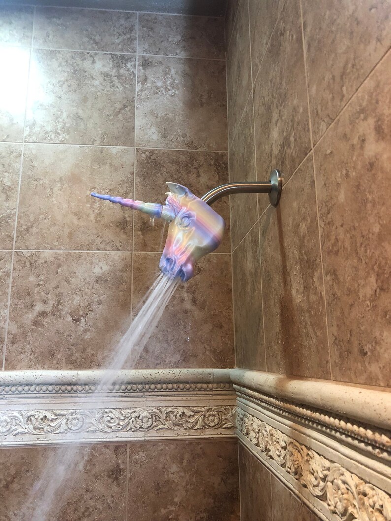 May include: A unicorn shower head with a rainbow design. The shower head is attached to a silver shower arm and is spraying water.