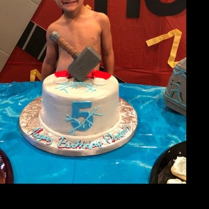 May include: A white birthday cake with blue frosting and a blue number 5 on top. The cake is decorated with blue frosting that looks like ice crystals. A toy hammer is on top of the cake. The cake is on a silver cake stand with the words "Happy Birthday Phoenix" written on it.