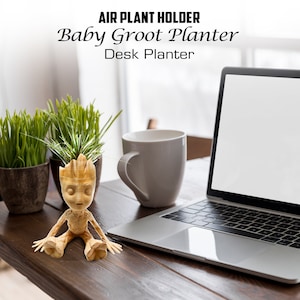 May include: A wooden Baby Groot planter with a small green air plant growing from its head. The planter is sitting on a wooden table next to a laptop and a white mug.