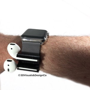 K&ouml;nnte beinhalten: Ein schwarz-wei&szlig;es 3D-gedrucktes Zubeh&ouml;r f&uuml;r eine Smartwatch, das zum Halten von kabellosen Ohrh&ouml;rern entwickelt wurde. Das Zubeh&ouml;r ist an einem silbernen Smartwatch-Armband mit einem schwarzen Mesh-Armband befestigt. Der Text "&copy;3DVisuals&DesignCo" ist im Bild sichtbar.
