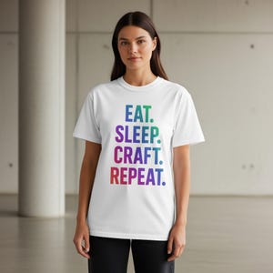 May include: White t-shirt with the words "EAT. SLEEP. CRAFT. REPEAT." in a gradient of blue, green, purple, and red. The shirt is a crew neck style and is being worn.