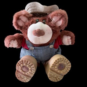 Puede incluir: Un oso de peluche con sombrero de paja, camisa roja y peto a rayas azules y blancas. El oso tiene una cara blanca con una nariz marrón y mejillas rosadas. El oso lleva botas color canela.