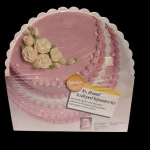 Könnte beinhalten: Ein rosa Tortentrenner-Set mit einem gewellten Rand. Das Set enthält eine runde 25,4 cm Platte mit weißem Zuckerguss und cremefarbenen Rosen. Die Verpackung enthält den Text "Wilton 10in Round Scalloped Separator Set".