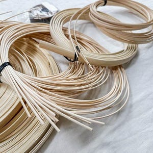 May include: Several bundles of natural-toned rattan cane and strips, some coiled and others loose. Black tape secures some bundles. The materials are displayed on a white surface, likely for craft projects or basketry.