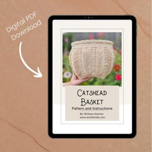 Könnte beinhalten: Digitaler PDF-Download auf einem Tablet angezeigt. Das Bild zeigt einen gewebten Korb in einer Hand, mit dem Text "Catshead Basket" und Anweisungen. Der Hintergrund ist eine verschwommene Szene im Freien.